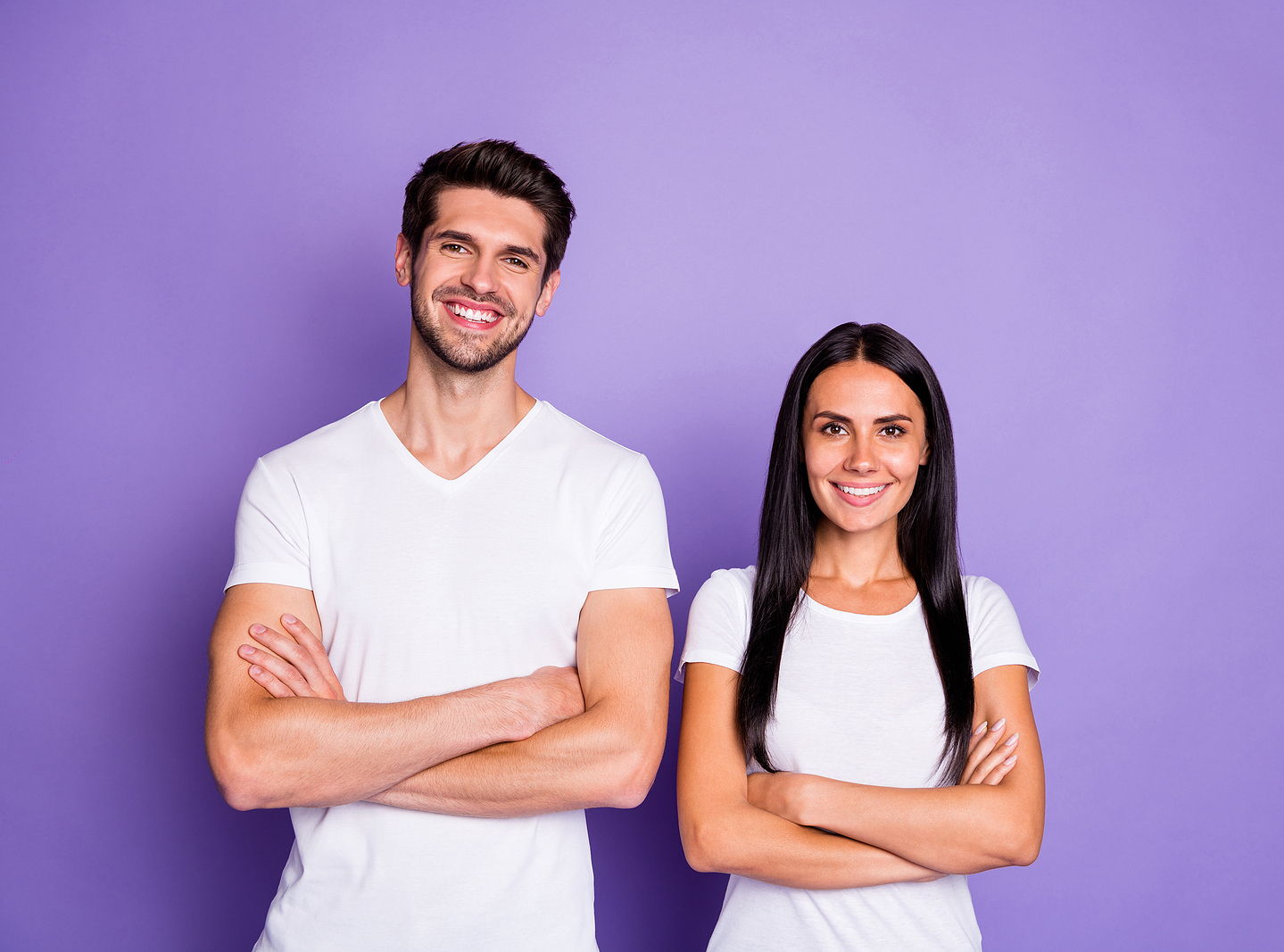 Image of an attractive man and woman of mixed ethnicities standing side by side with their arms crossed and a white tshirt, smiling against a purple background.