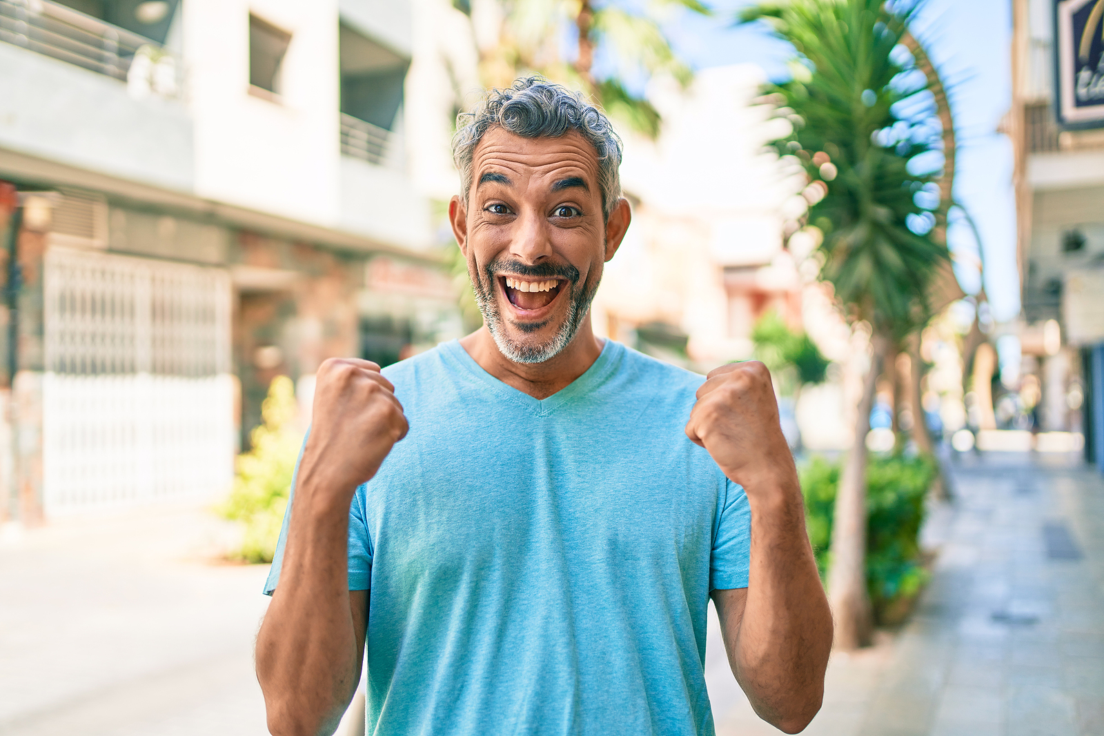 An attractive older man with white hair and a beard laughs while holding his arms in fists confidently while at a plaza.