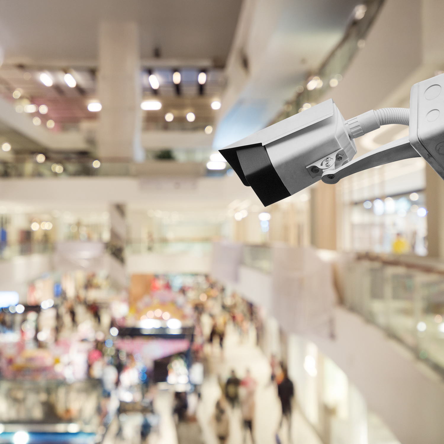 A security camera in focus overlooks a busy, brightly lit shopping mall with blurred crowds and multiple levels in the background.