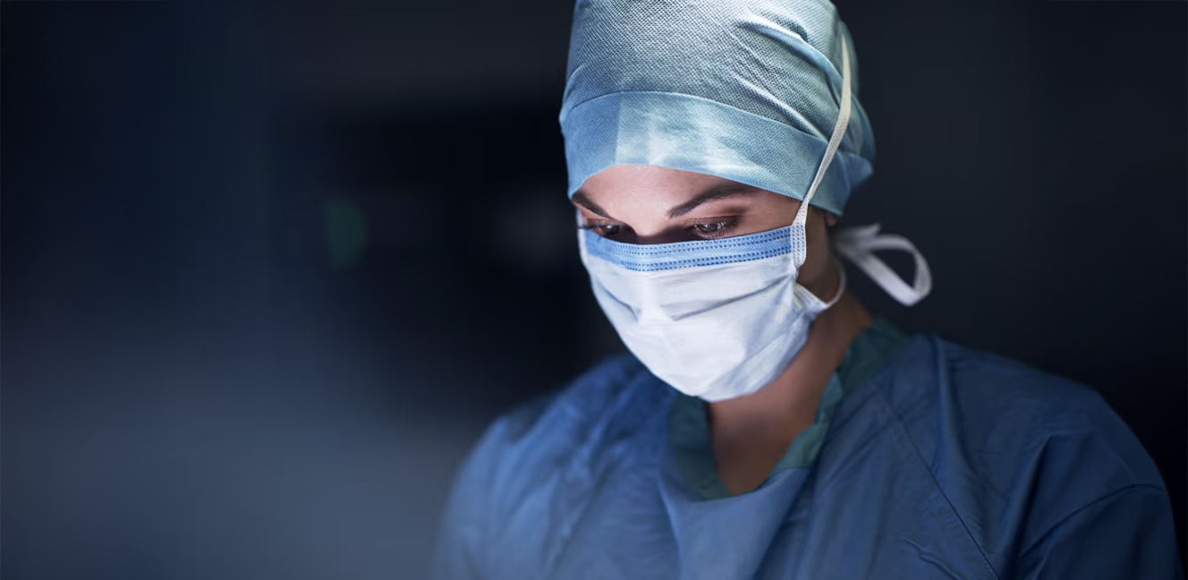A healthcare worker wearing a surgical mask, cap, and scrubs looks down in a focused manner against a dark background.