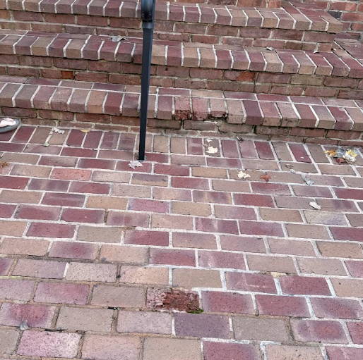 Brick walkway and steps with visible cracking, loose bricks, and deteriorating mortar joints near a metal handrail.