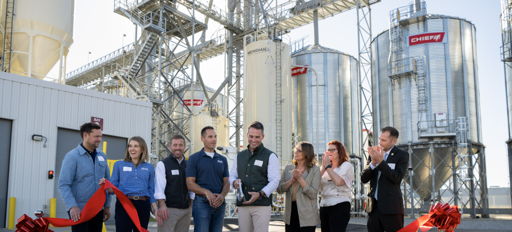 Group smiles at a ribbon-cutting ceremony for a new facility, with silos in the background, highlighting community and celebration.