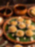 Small Indian sweets in paper liners, topped with cashews and pistachio crumbs on a banana leaf–lined clay plate, with earthen pots and scattered nuts in the background.