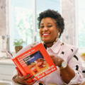 A person holds a reddish orange book titled 'Watermelon and Red Birds' in a brightly lit kitchen