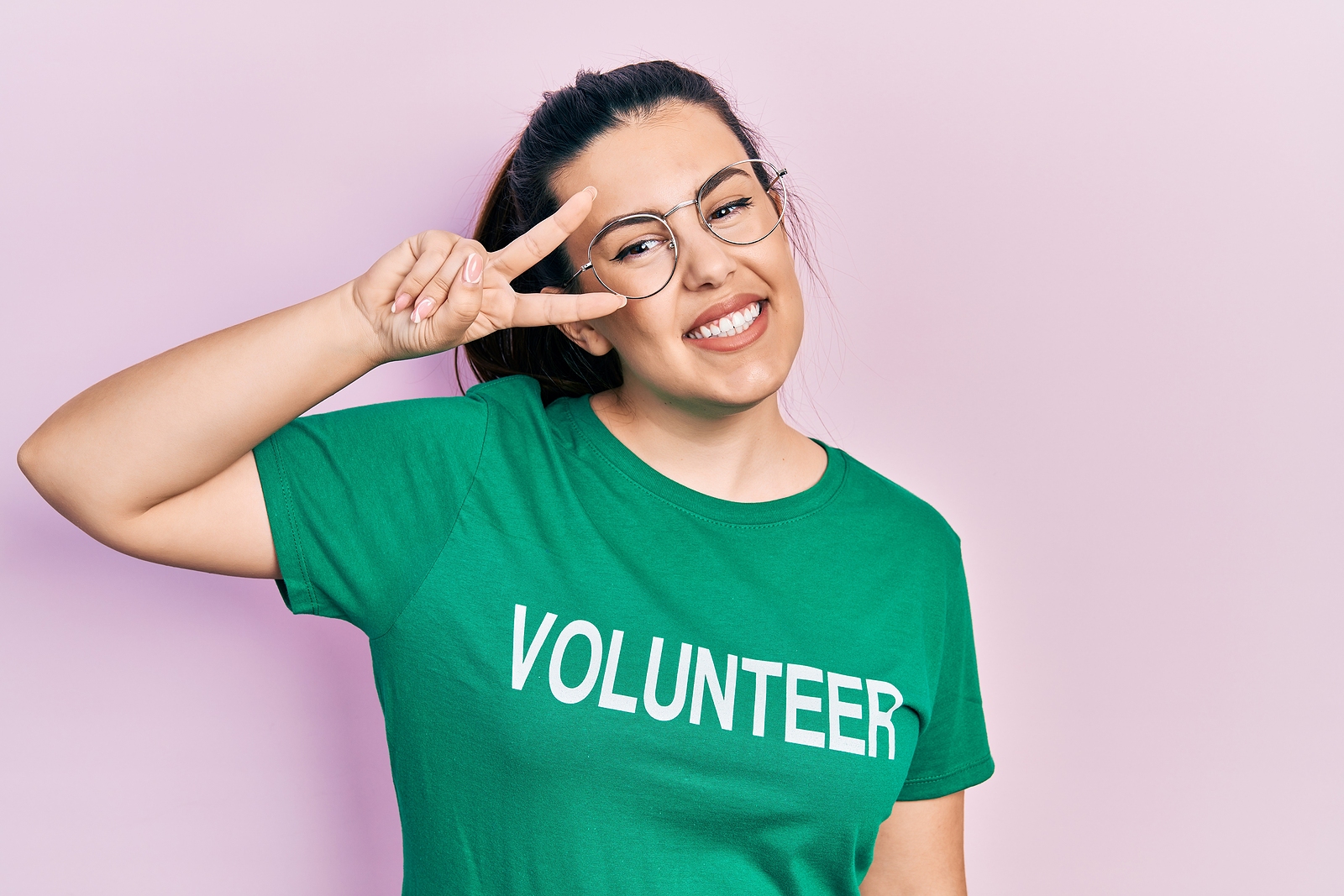 Young hispanic woman wearing volunteer t shirt doing peace symbol with fingers over face, smiling cheerful showing victory.