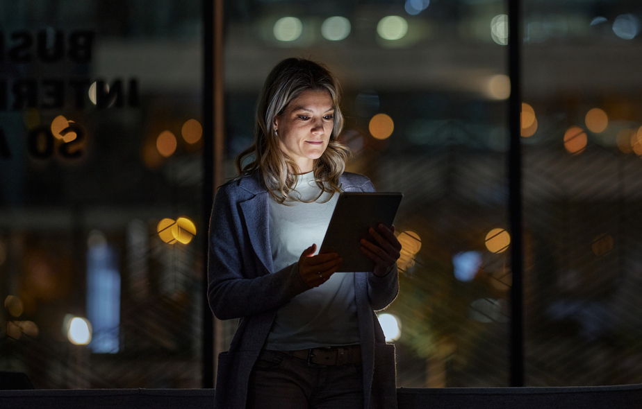 A woman stands indoors at night, illuminated by the glow of a tablet in her hands, with blurred city lights visible through large windows behind her.