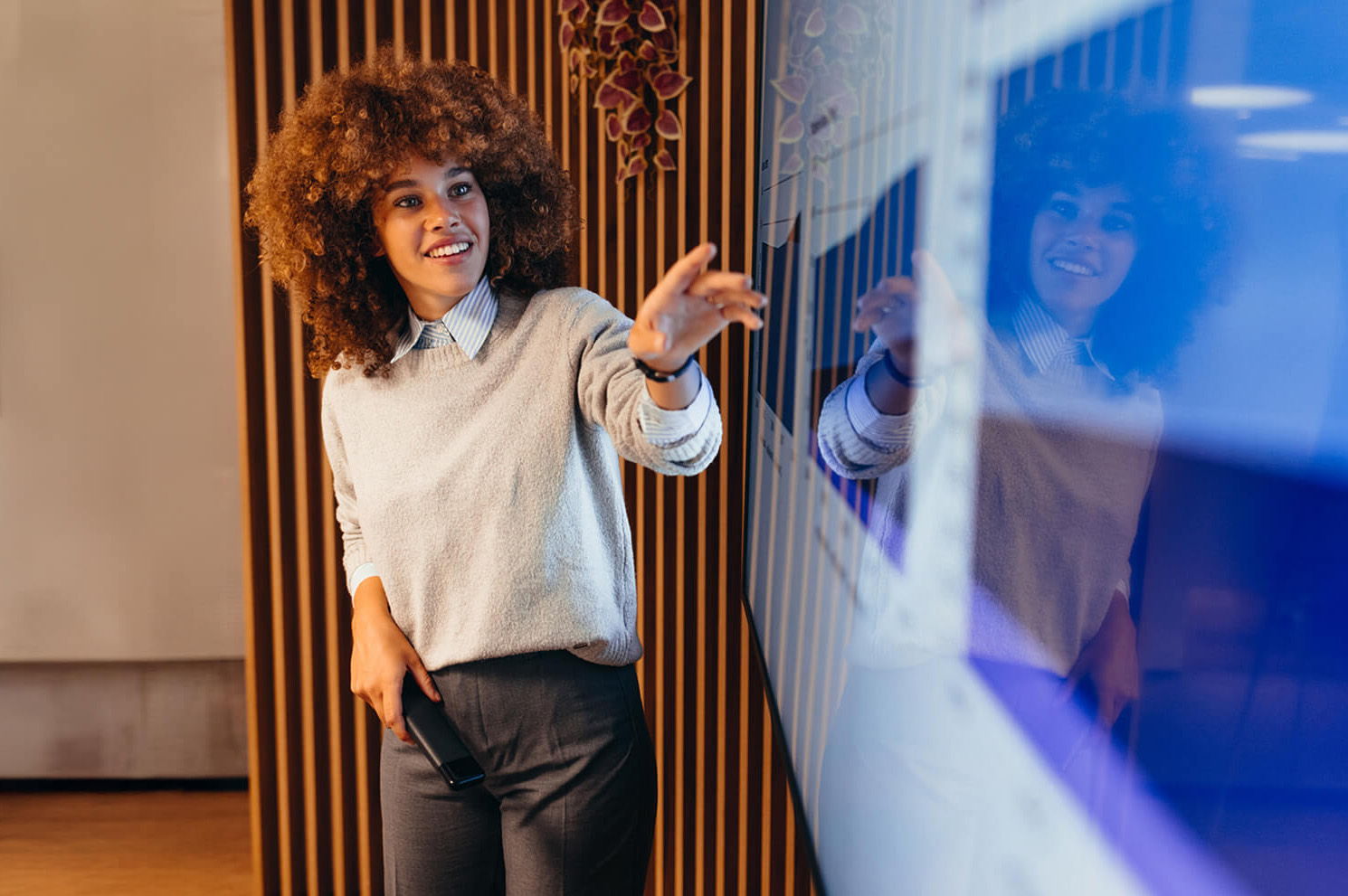A woman with curly hair smiles and gestures toward a large screen during a presentation. She holds a remote control and stands in a modern, well-lit office space with wooden paneling behind her.