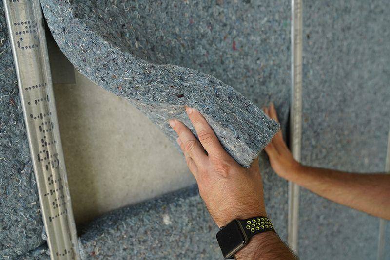 A person wearing a smartwatch installs a blue-gray panel of recycled insulation material onto a wall, lifting the flexible panel to fit it into a metal frame.