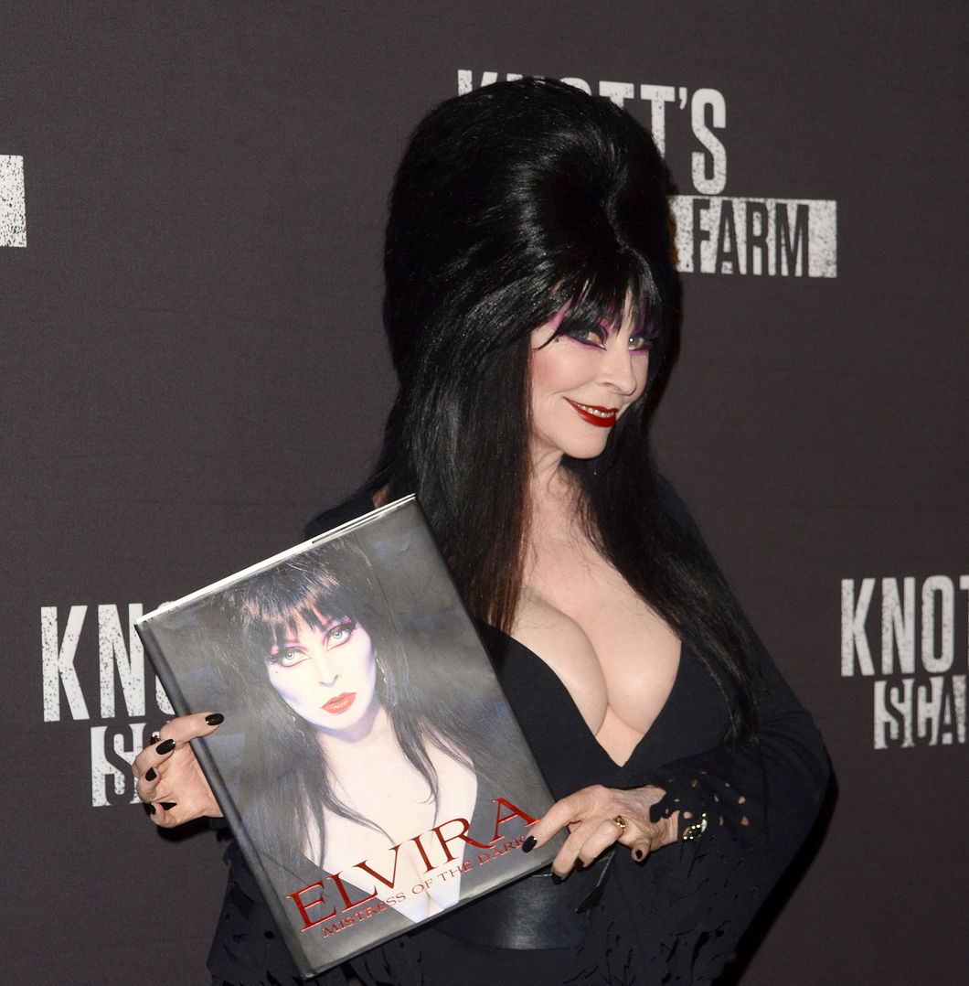 Elvira at the 2016 Knott's Scary Farm at Knott's Berry Farm  holding her book and smiling.