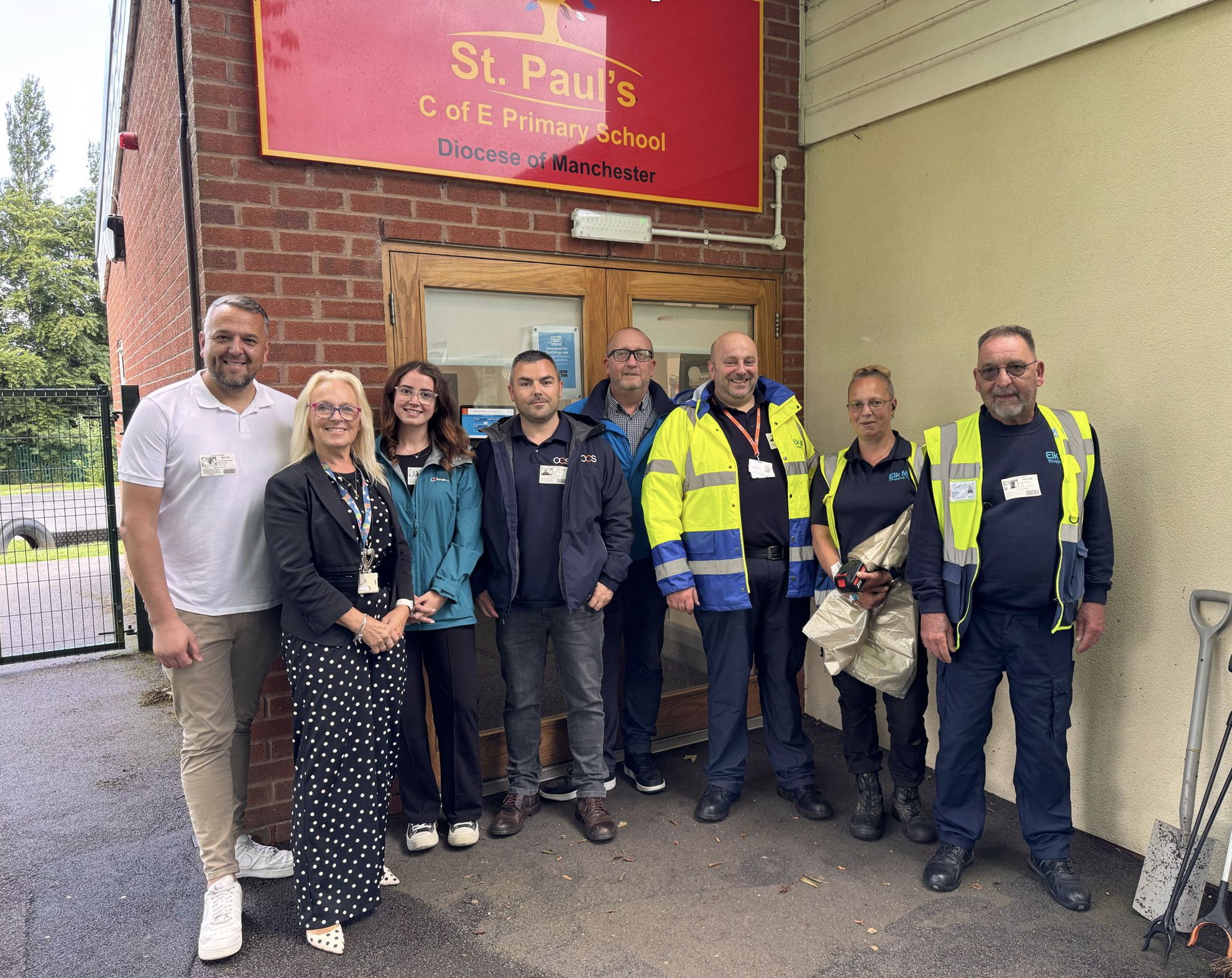 A group of eight adults, some in high-visibility jackets, stand smiling in front of the entrance to St. Pauls C of E Primary School, beneath a red sign, outdoors on a paved area.
