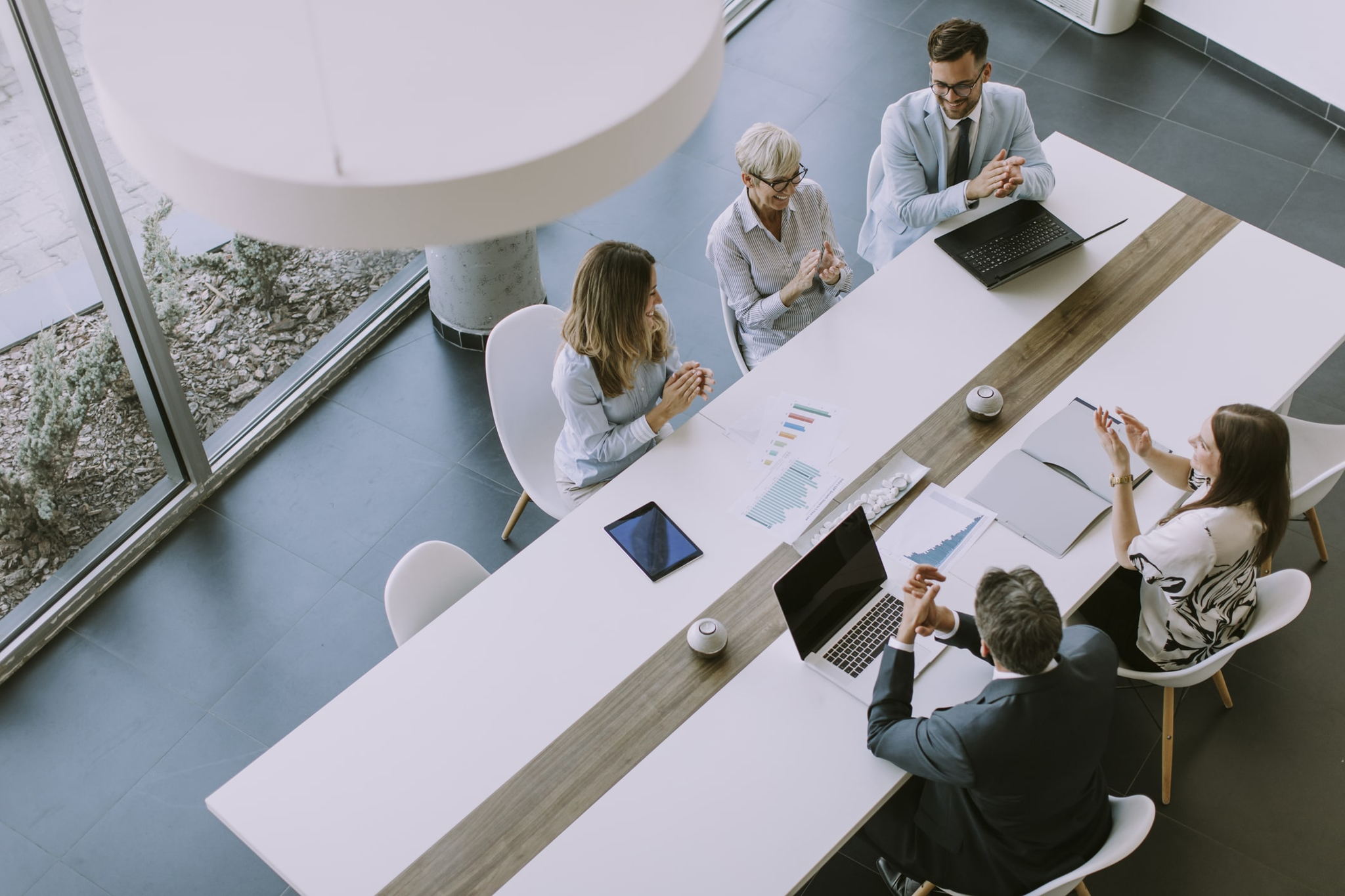 Five people in a modern office setting, designed through our workplace services, are seated around a long white table with laptops and documents. They appear to be engaged in a meeting, with some people using gestures. Large windows provide natural light.