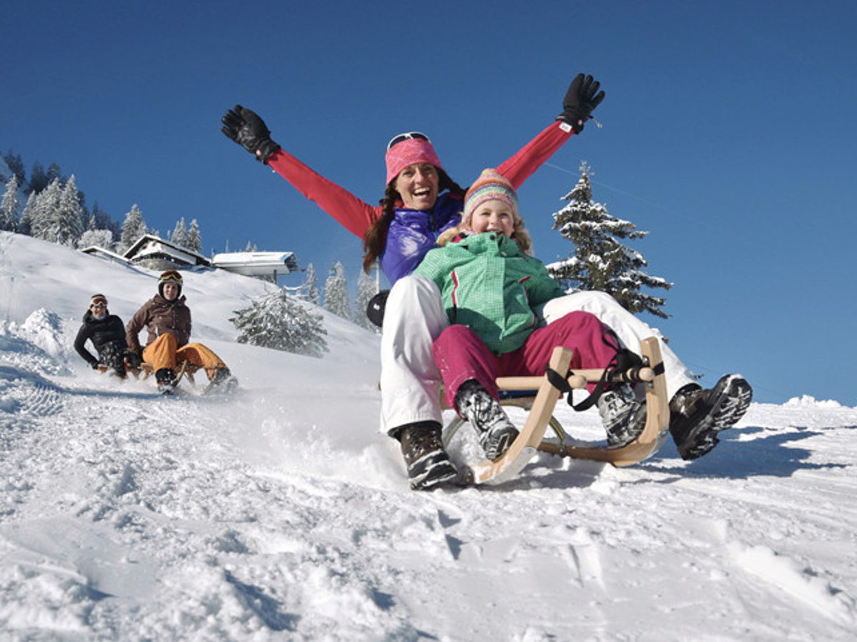 Mutter mit Kind auf dem Schoß fährt mit Holzschlitten schneebedeckten Hügel herunter. Die Mutter hält Jubelnd die Arme hoch. In Distanz zwei weitere Personen, die auf einem Holzschlitten folgen.