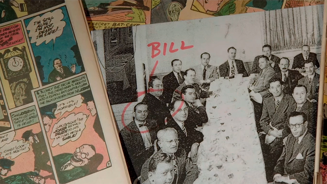 Image from the documentary of a black and white image of Bill sitting at a table with others and the Batman comics in the background.