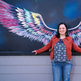 A smiling woman in casual attire stands before a colorful wing mural, creating a joyful and uplifting atmosphere.