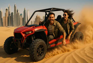 Excited couple riding a red dune buggy fast through the desert, kicking up sand with a modern city skyline visible in the distance under a clear sky.