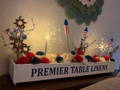 4th of july gome decorations with rockets, lights, and flowers on a wood piece with premier table linens written on it