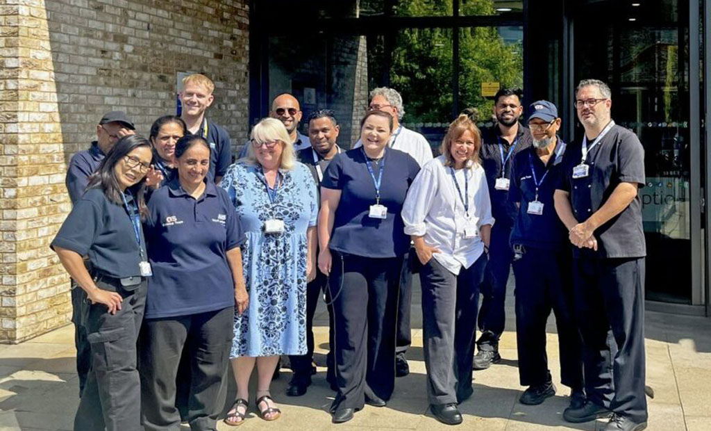 A group of thirteen people, some in uniforms and others in casual clothing, stand smiling together outside a building with glass doors and a brick wall on a sunny day.
