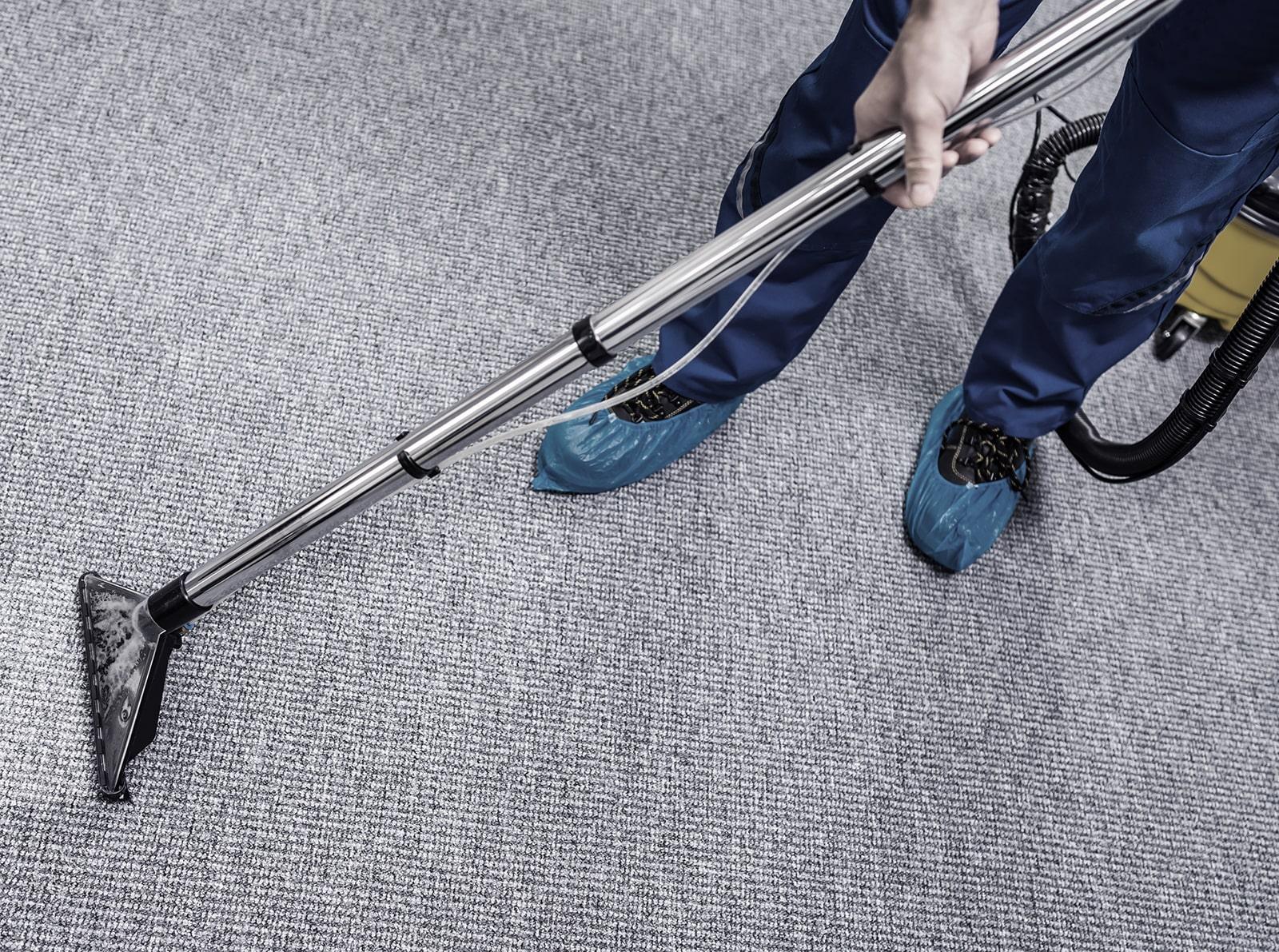 A person wearing blue shoe covers is cleaning a gray carpet with a vacuum cleaner, viewed from above. The vacuums long handle and nozzle are visible as they move across the carpet’s textured surface.