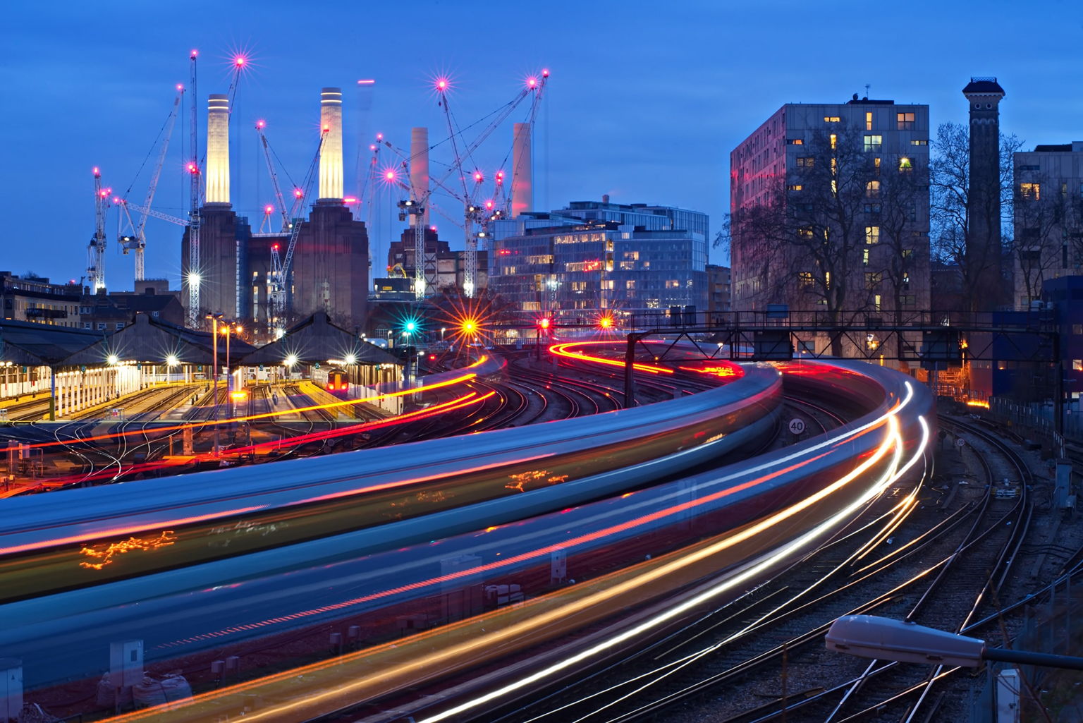 Cityscape at dusk with curving train light trails, illuminated modern buildings, lit-up construction cranes, and four tall chimneys in the background. The sky is blue and the scene is vibrant with urban activity.
