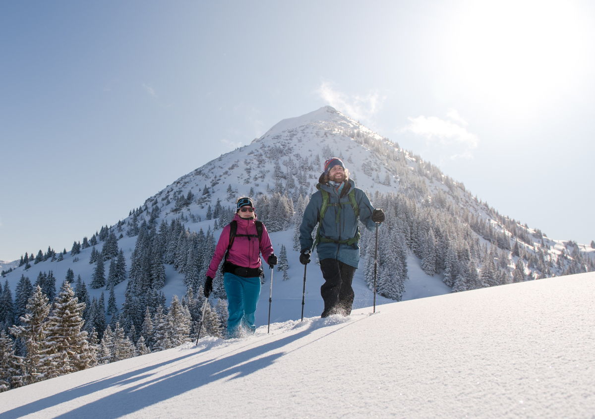 Zwei Schneeschuhwanderer laufen mit Stöcken im Pulverschnee. Sonniges Wetter mit Bergspitze mittig im Hintergrund.