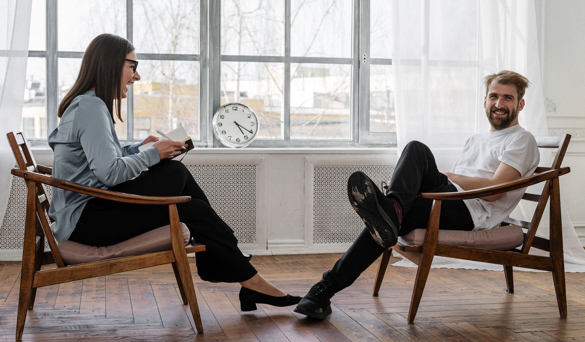 A man sitting in front of a therapist. She is laughing and he is looking at the camera and laughing as well.