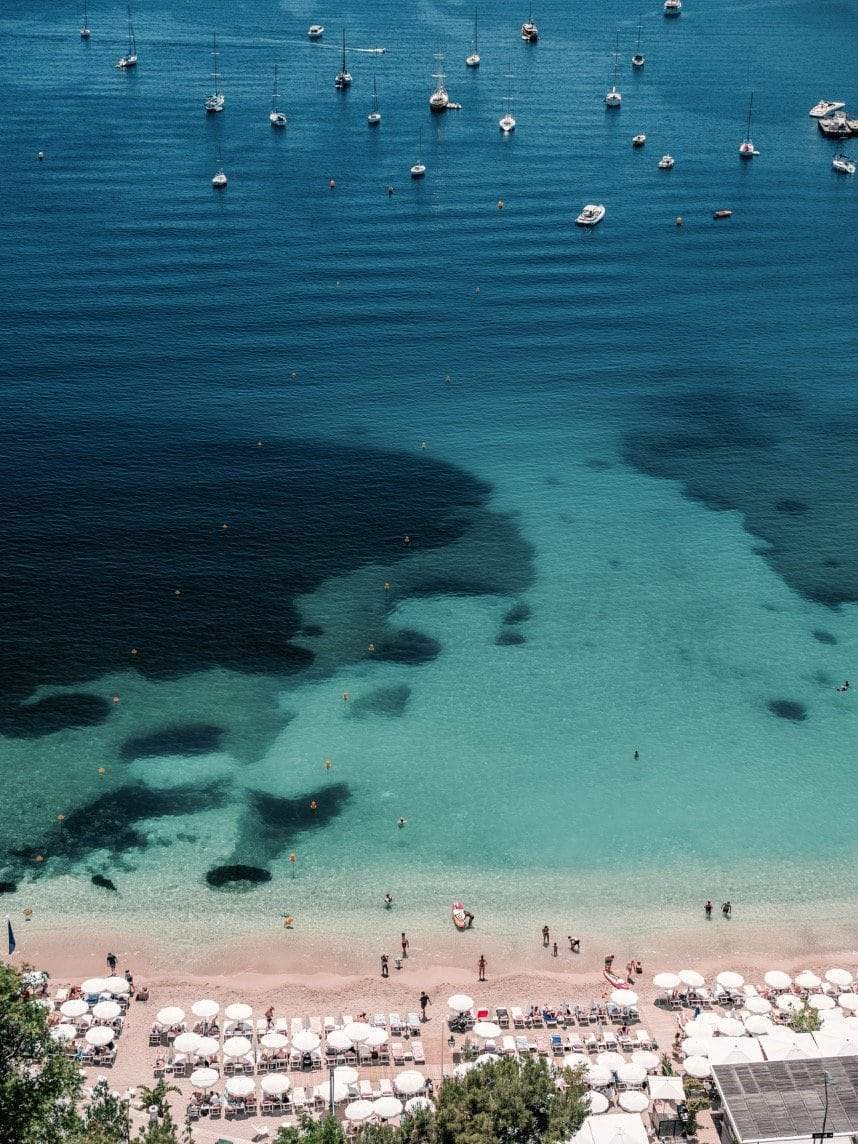 Villefranche-sur-Mer by Stuart Cantor - A coastal photograph of the blue and green waters of Villefranche-sur-Mer with boats moored in the ocean and holiday-makers on the beach.