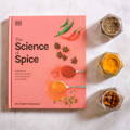 A light pink book titled 'The Science of Spice' lays on a countertop next to three open spice jars of differing contents 