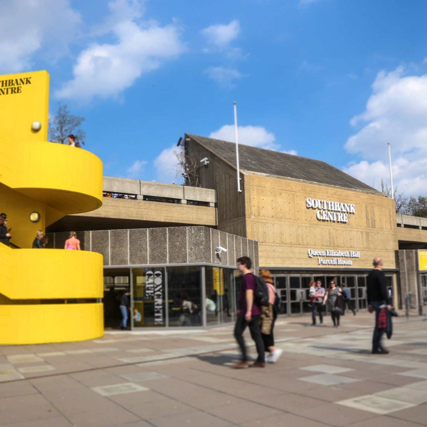 People walk outside the Southbank Centre in London on a sunny day. The building is modern with concrete and yellow accents, and the Southbank Centre sign is visible above the entrance.