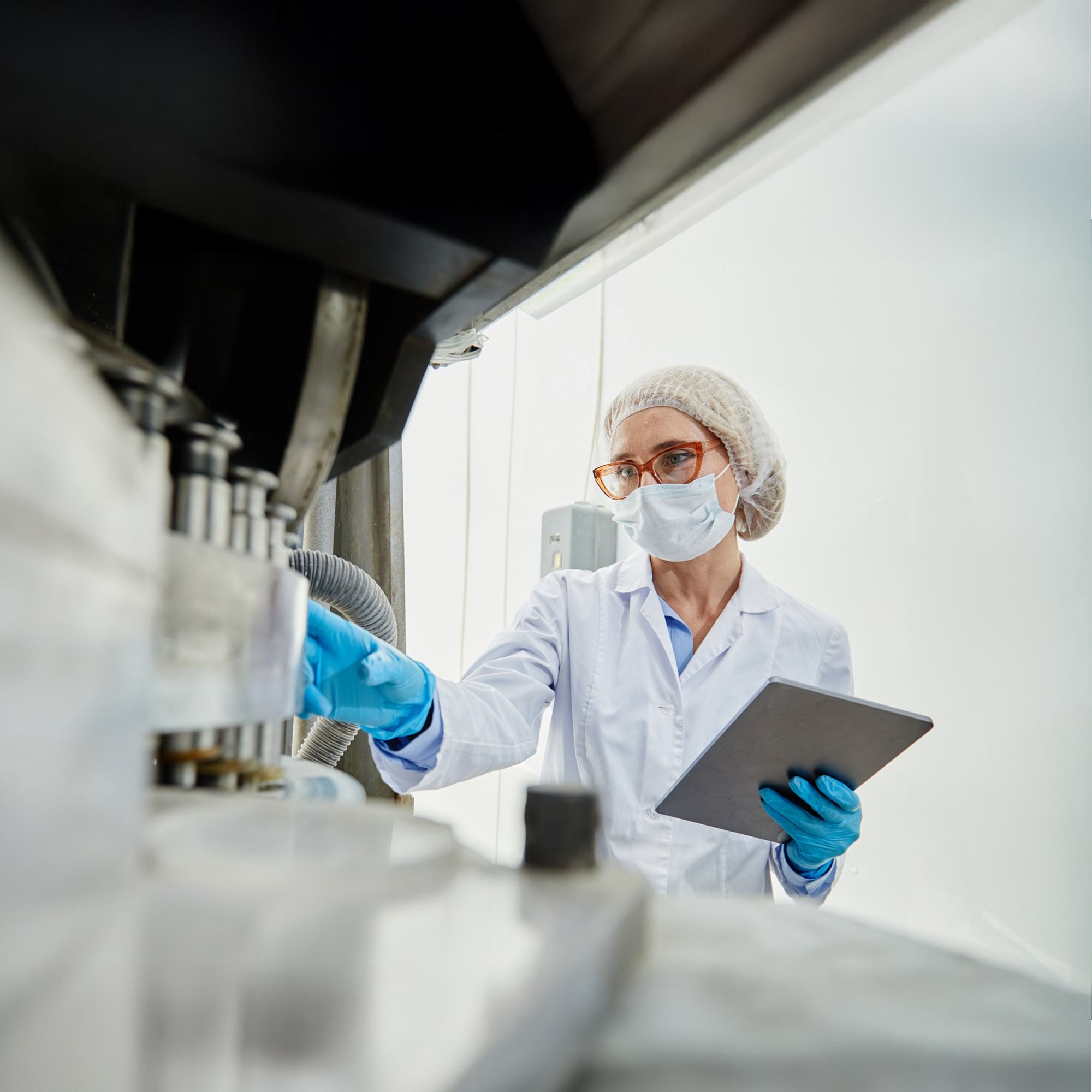 A scientist wearing a lab coat, hairnet, mask, and gloves operates laboratory equipment while holding a tablet, working in a clean, bright lab environment.