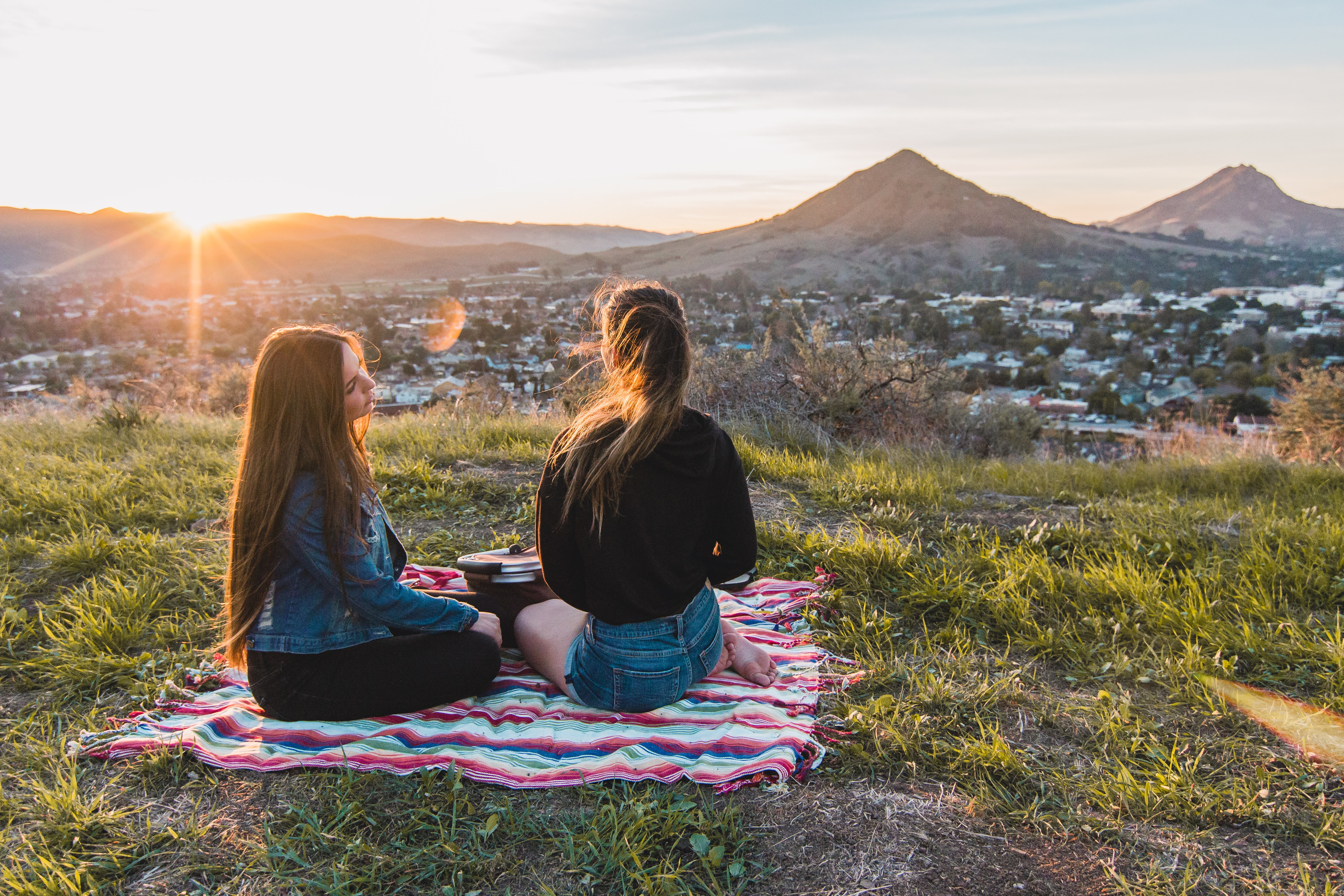 Two women sit together on a blanket at a hill overlooking their town during a sunrise.