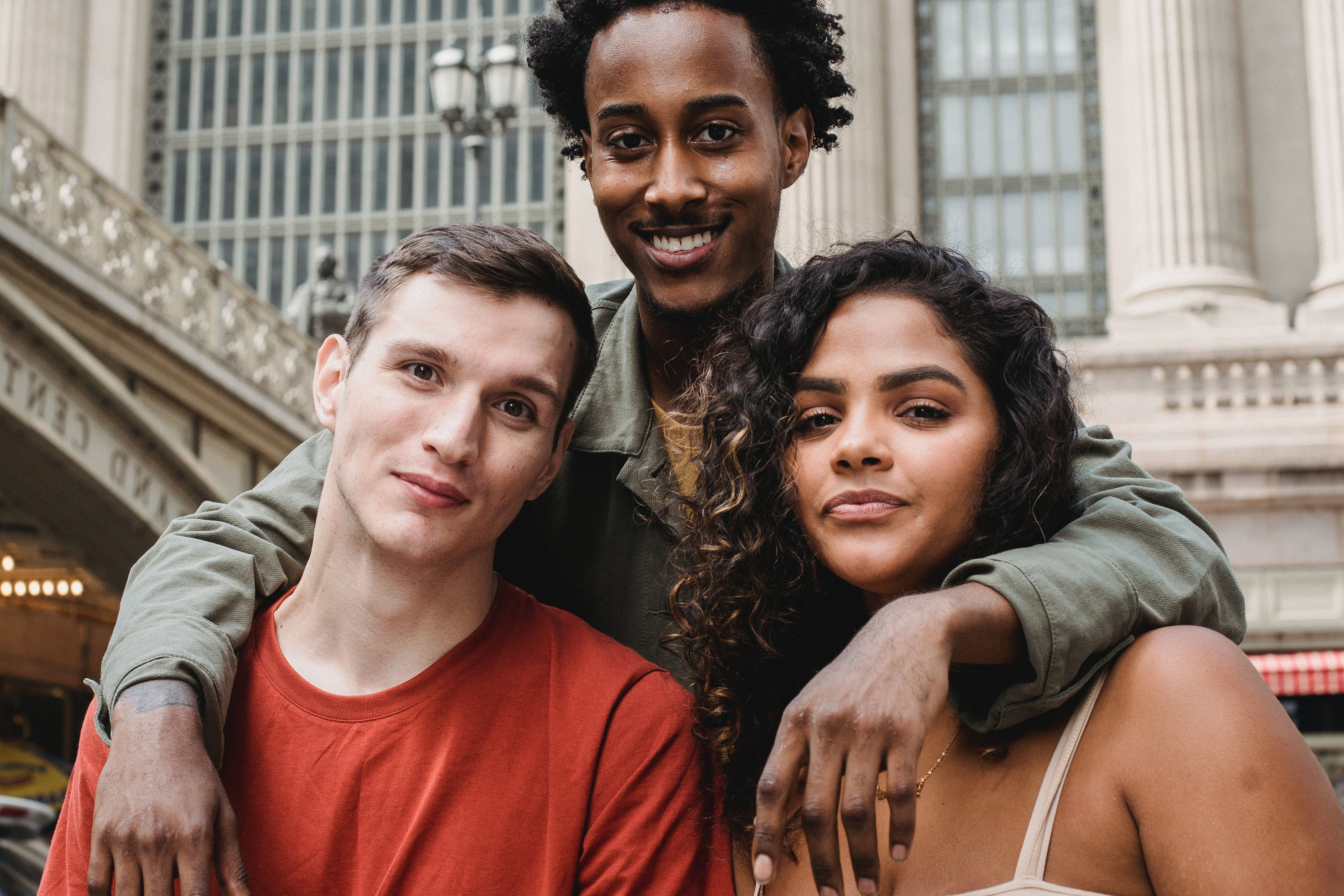 Image of 3 multi ethnic attractive friends. There is a man with a red shirt and a woman with curly hair being hugged by another friend above them.