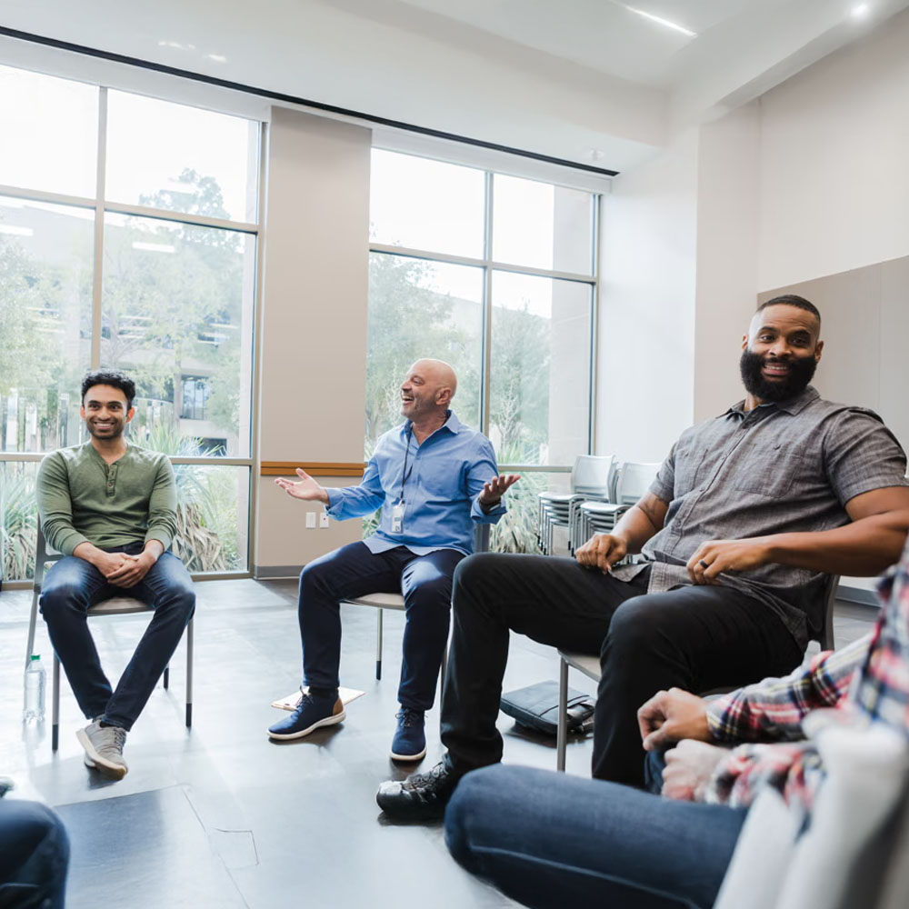 Four men sit in a bright room with large windows, arranged in a circular formation, smiling and engaged in conversation, suggesting a group discussion or support meeting.