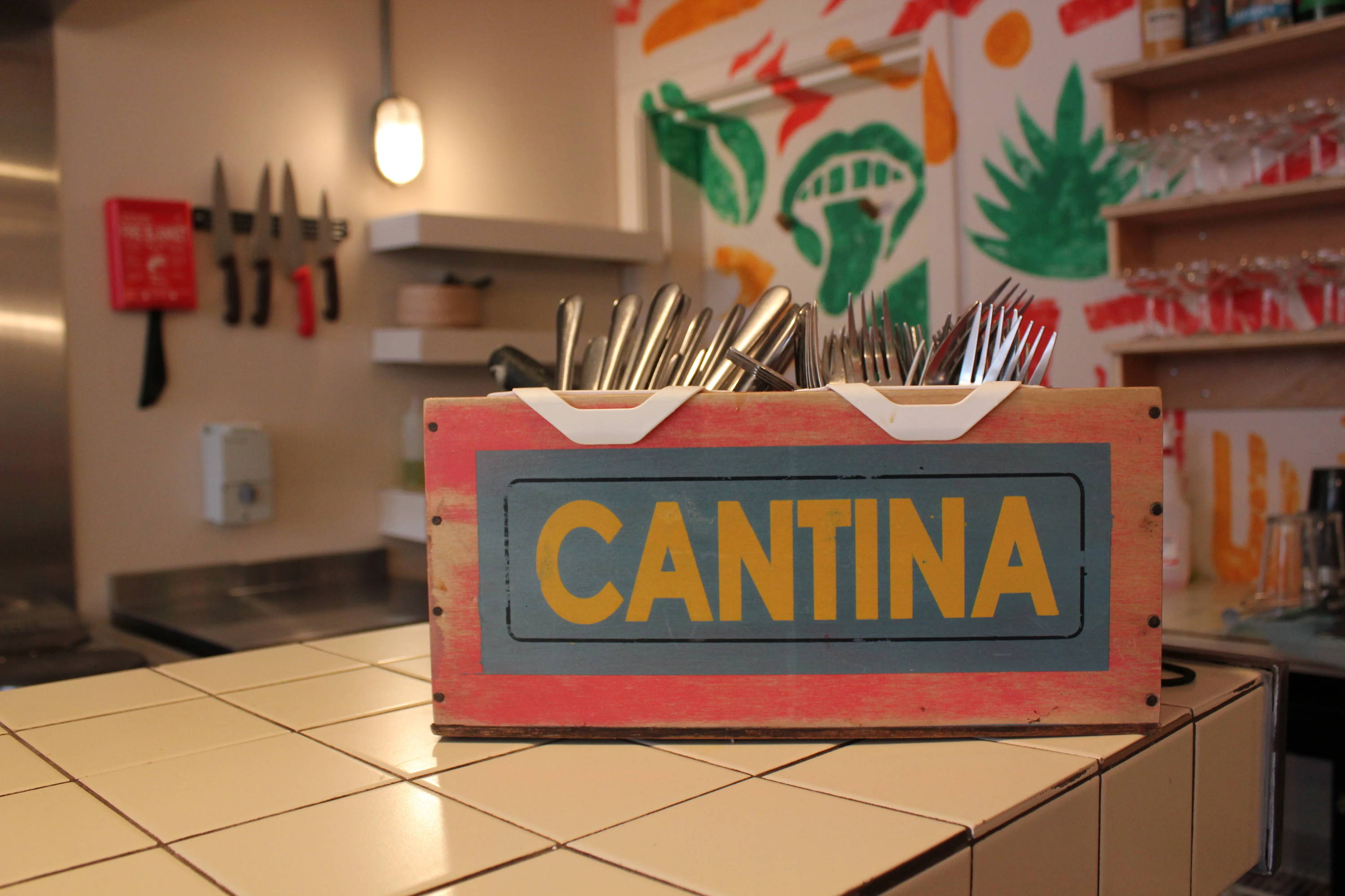 A red and blue rustic wooden pot which is holding stainless steel knives and forks. The box is placed on a white squaretiled bar.