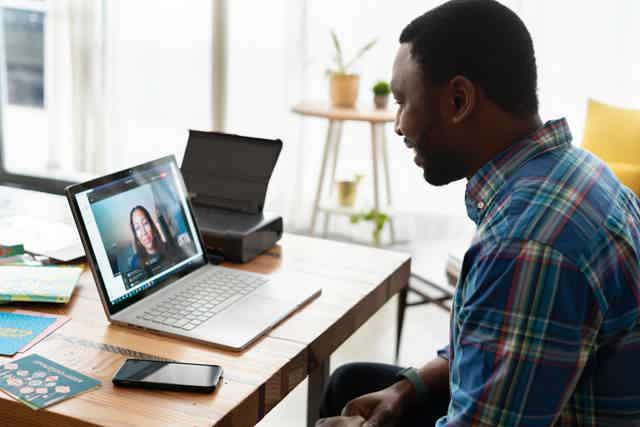 Man in plaid shirt on video call with woman on laptop screen; workspace with phone, printer, plants.