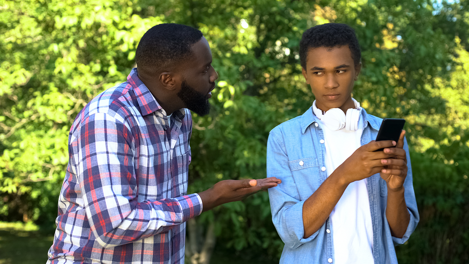 A black father scolding his teenage son who is on his phone giving the side eye.