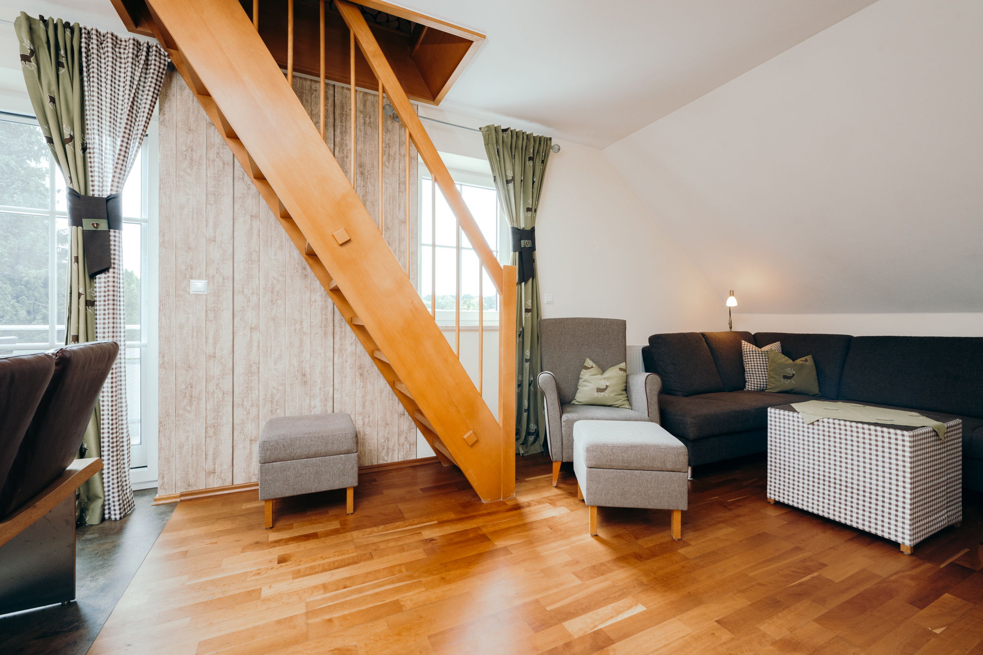 Ein Wohnzimmer mit Holzboden, Sofa, Sessel und einer hölzernen Treppe in das Obergeschoss.