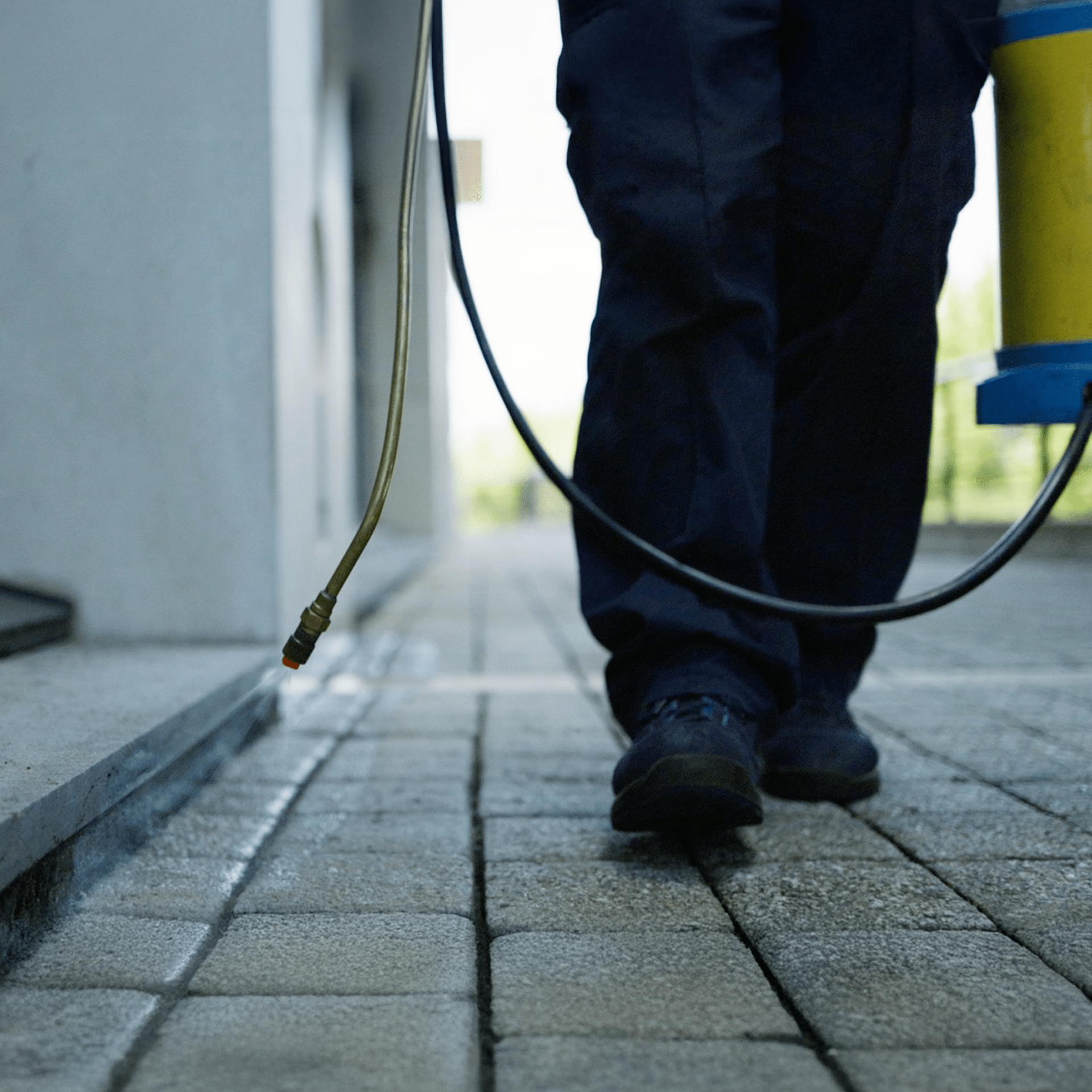 A person in dark pants and shoes sprays a paved walkway with a yellow and blue canister, likely applying weed killer or pesticide outdoors near a building.