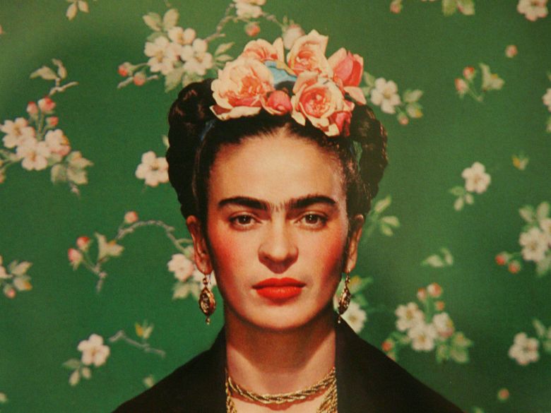 Frida drawn realistically, with flowers in her braided hair and red lipstick. She is in front of a wall with rose wallpaper.