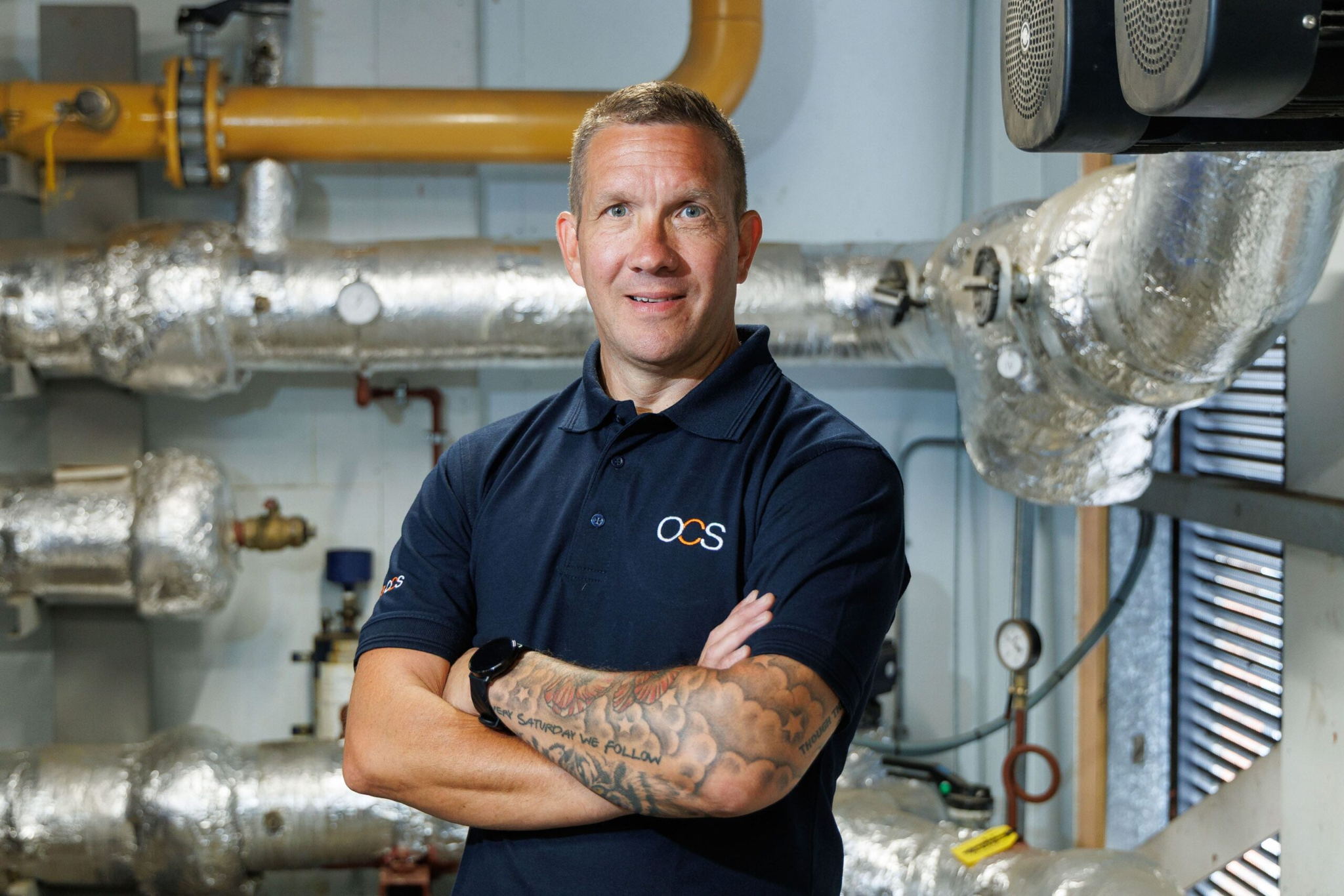 A man in a navy OCS polo shirt, with tattooed forearms, stands confidently with arms crossed in front of industrial pipes and equipment wrapped in insulation.