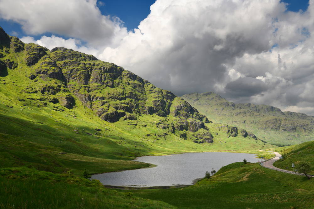 A mountain loch at the famous Rest and be Thankful pass, surrounded by lush grass and a steep mountain cliff on one side.