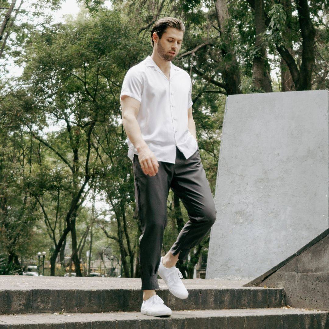 model walking down some steps in a park wearing white shoes grey pants and an untucked short sleeve white silk shirt from 1000 kingdoms