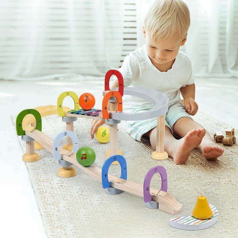 Un tout-petit joue avec un jouet en bois coloré InvenToy Montessori Musical Marble Run sur un sol recouvert de moquette dans une pièce lumineuse et ensoleillée.