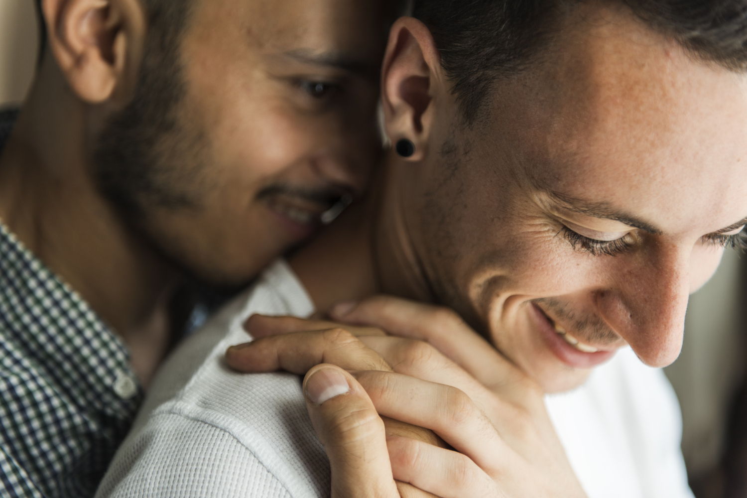 A close up of a male couple having an intimate moment. One is hugging the other from behind him.
