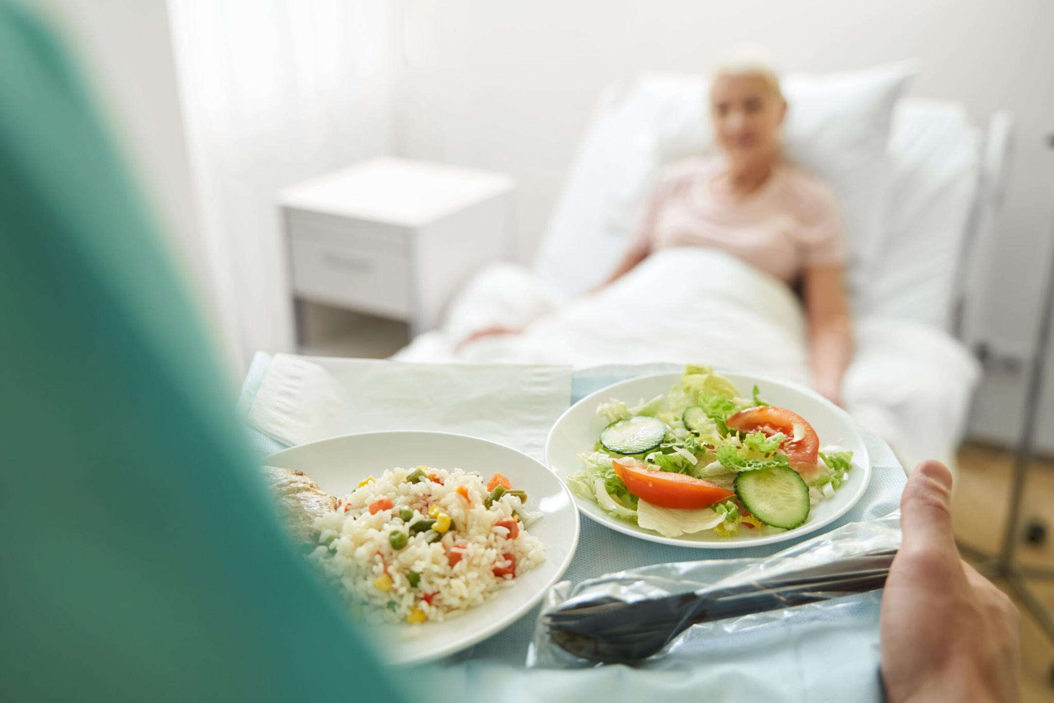A person in a hospital bed is blurred in the background while a tray with a plate of rice and vegetables, a fresh salad, and utensils is in focus in the foreground.
