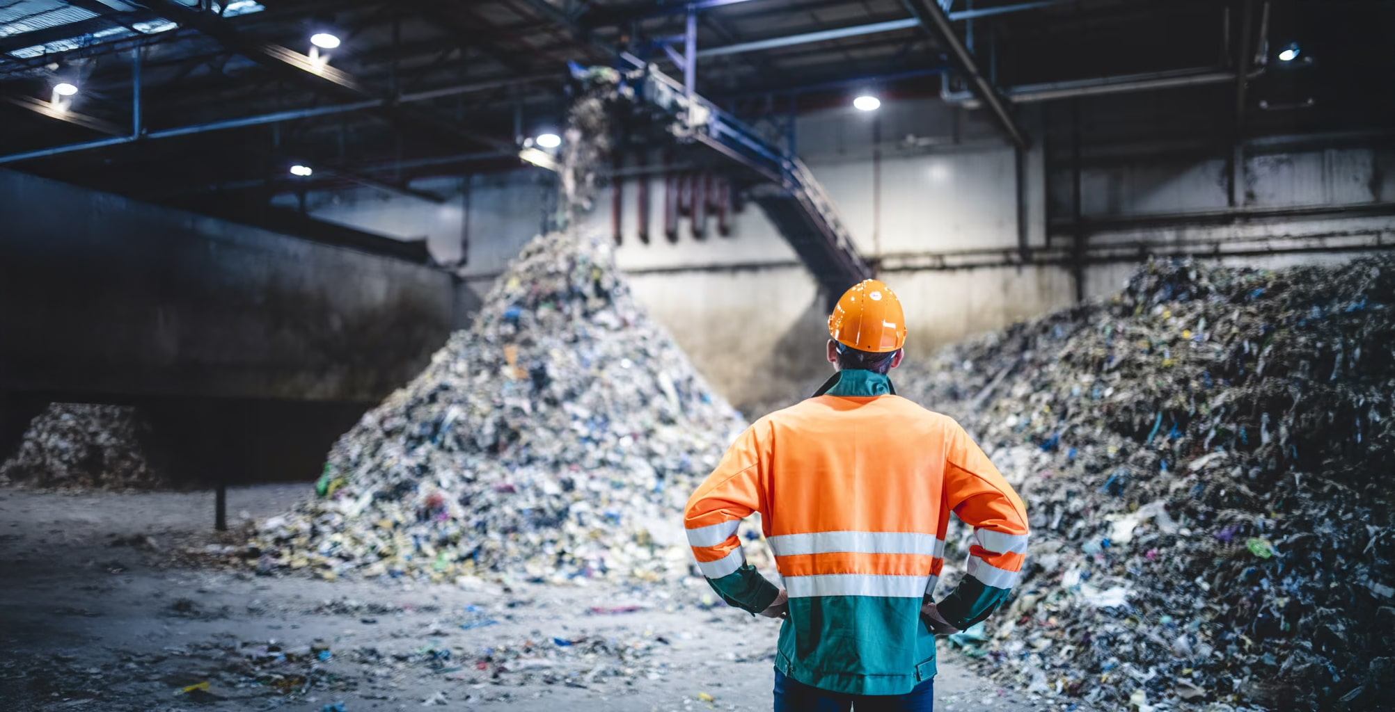 A worker in a high-visibility jacket and hard hat stands with hands on hips, facing large piles of waste inside an industrial recycling or waste processing facility.