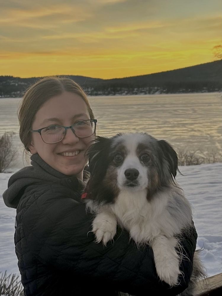 A young white woman wearing glasses is smiling while holding a small black and white dog. She is standing in front of a snowy lake at sunset.