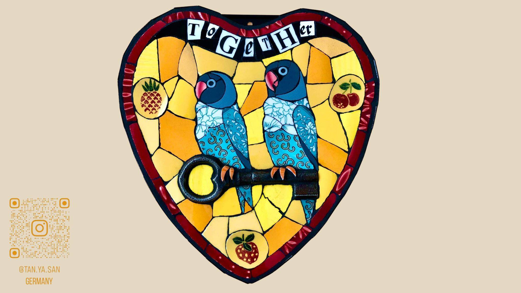 Twee blauwe papegaaien zittend op een sleutel in een gele hartvorm met de woorden ‘Together’ bovenaan. Dit is gemaakt in mozaiek met oude porseleinen borden en kopjes in Picassiette stijl.