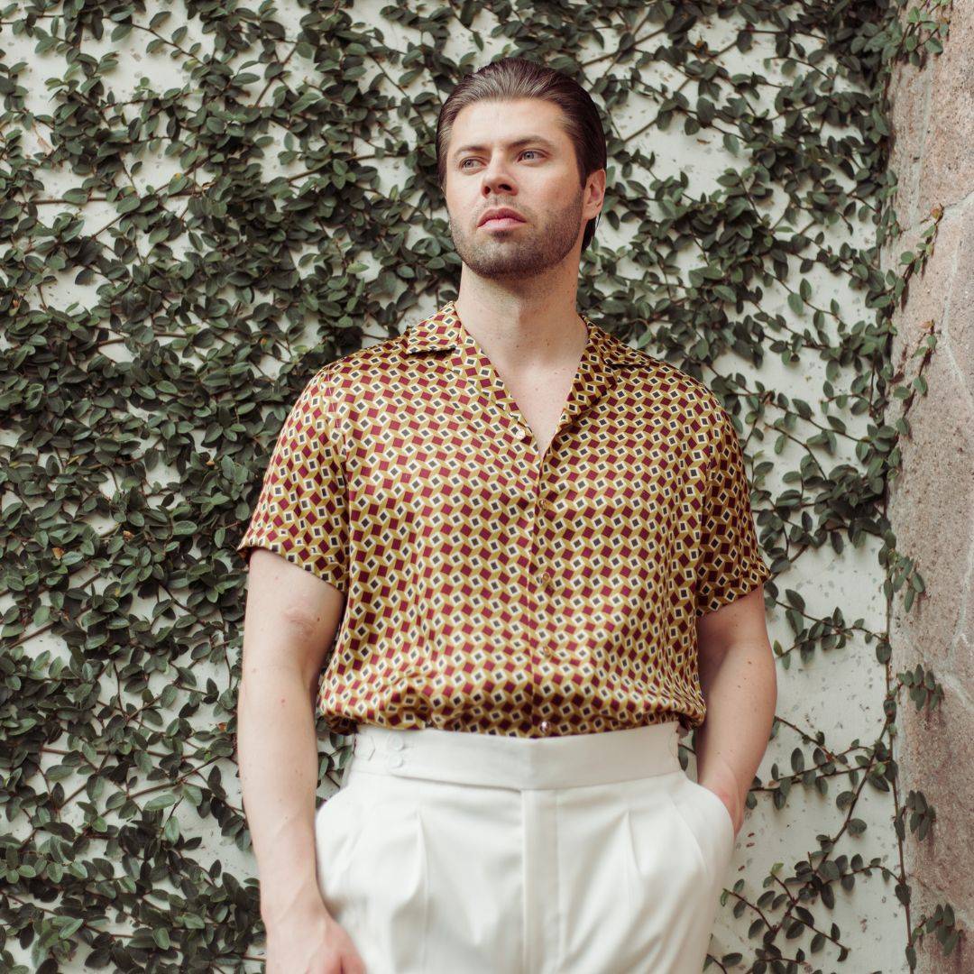 model standing in the garden and wearing a short sleeve gold bowling silk shirt from 1000 kingdoms with white pants