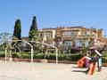 Sonniger Spielplatz mit Rutsche, Spielhaus und Fußballtoren in einem Sandbereich, vor einer Kulisse aus Bäumen und Gebäuden.
