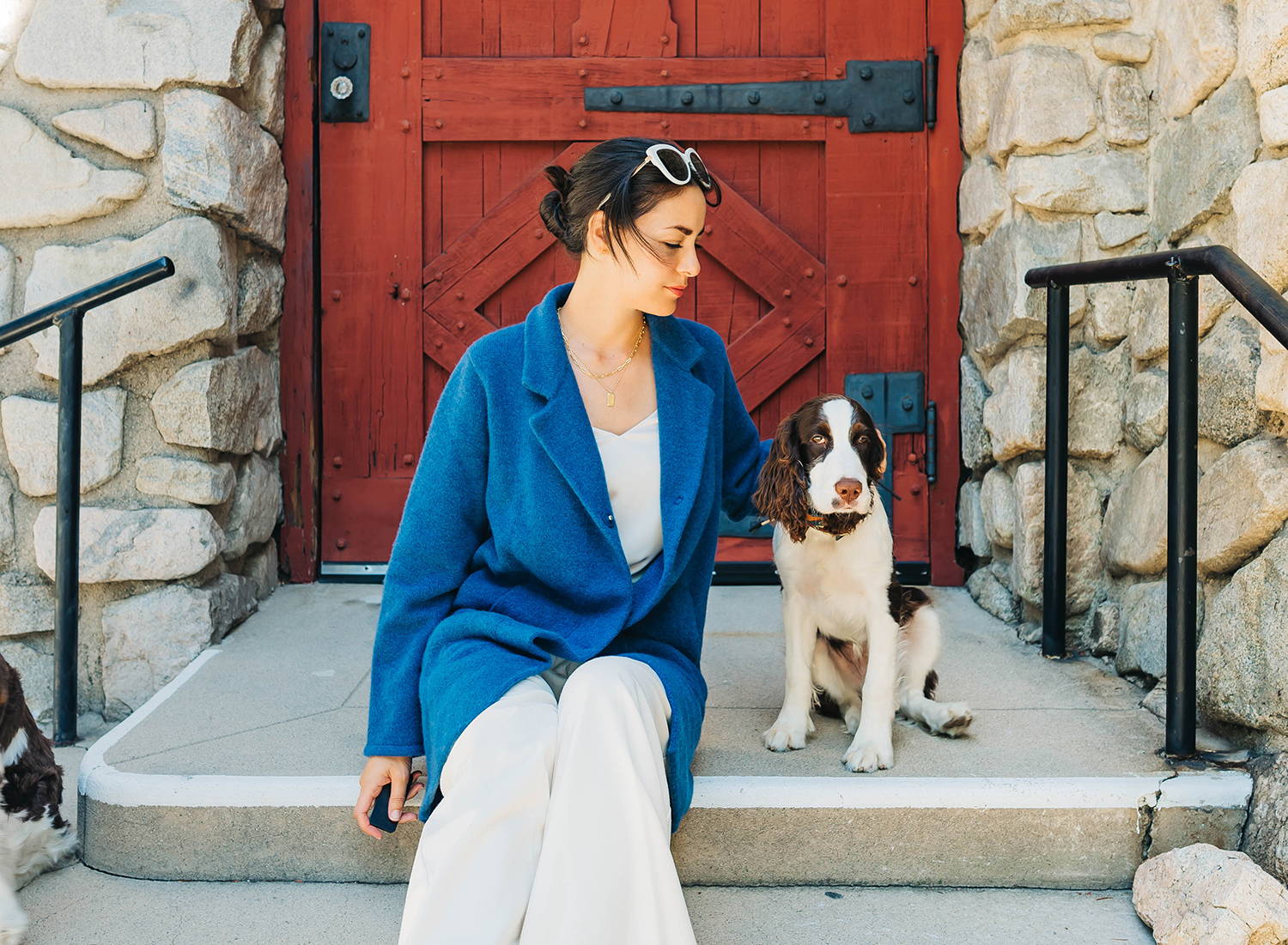 A woman wearing a blue coat, white v neck top and a pair of white pant sitting on the stair and petting her dog which sits next to her.